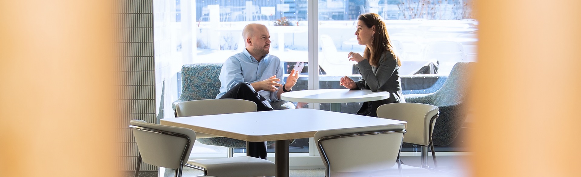 Two people sit at a small round table in a bright lounge area, talking near large windows that overlook the city.
