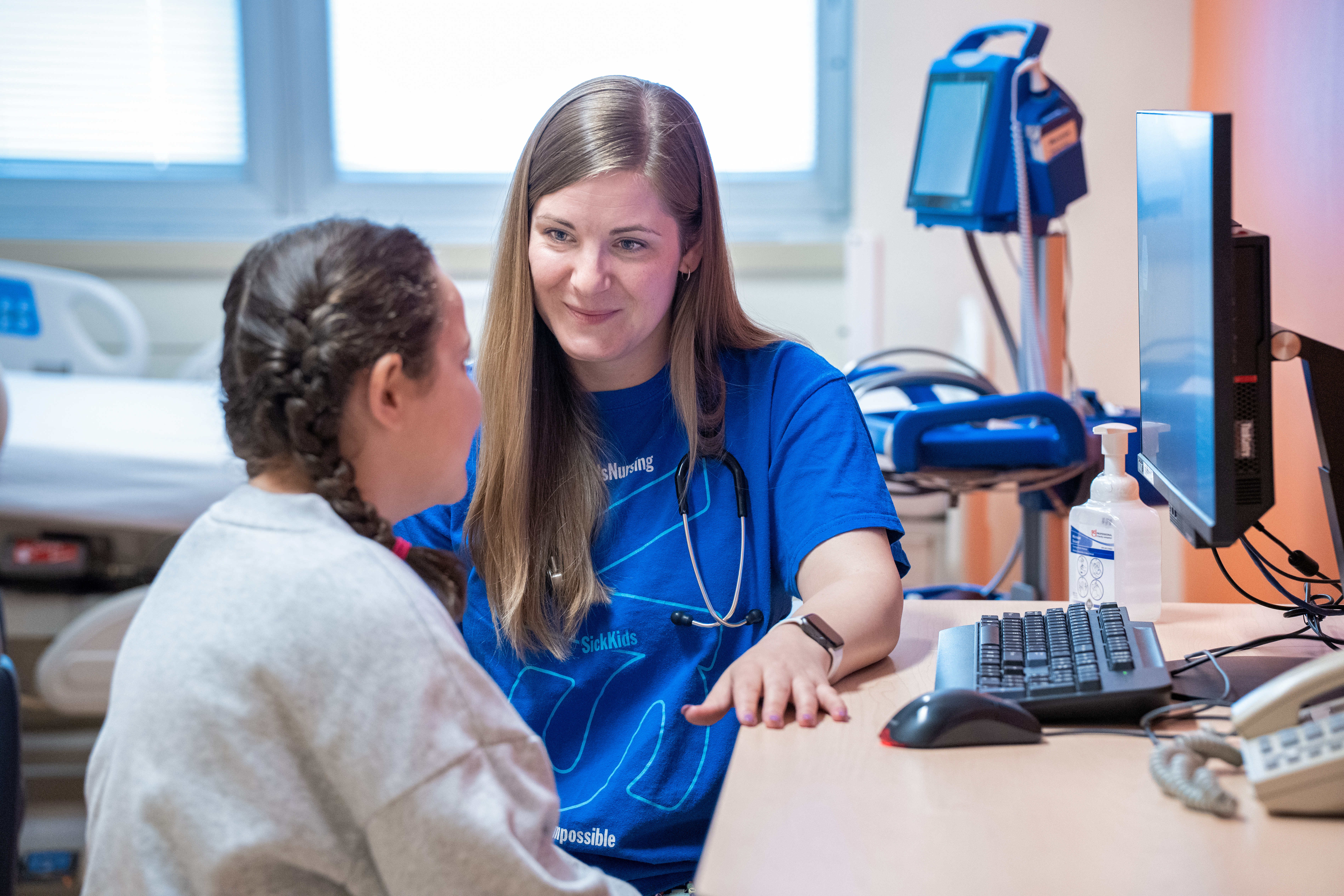 Healthcare worker in blue scrubs with stethoscope gently holding hands with a young patient in gray sweatshirt, providing comfort and care in a bright medical setting.RetryClaude can make mistakes. Please double-check responses.
